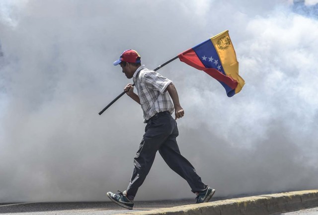 An opposition demonstrator carries the national flag in clashes with the riot police during a demonstration against Venezuelan President Nicolas Maduro in Caracas, on May 26, 2017. Riot police in Venezuela fired tear gas and water cannon to stop anti-government protesters from marching on a key military installation Friday during the latest violence in nearly two months of unrest. / AFP PHOTO / JUAN BARRETO